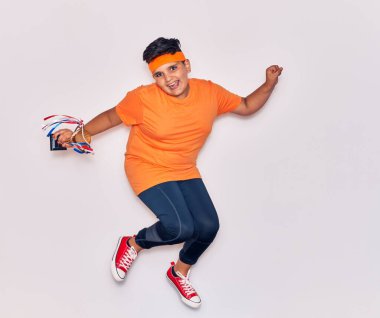 Spor kıyafetleri giyen sevimli İspanyol çocuk mutlu gülümsüyor. Yüzünde tebessümle zıplıyor İzole edilmiş beyaz arka planda ödül tutuyordu