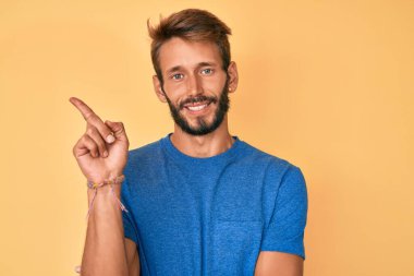 Sakallı, sıradan kıyafetler giyen, yüzünde kocaman bir gülümseme olan, eli ve parmağı kameraya bakan yakışıklı beyaz bir adam.. 