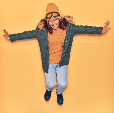 Kışlık giysiler giyen genç, güzel Latin kadın mutlu gülümsüyor. Yüzünde ve kollarında tebessümle, izole edilmiş sarı arka planda atlıyor.