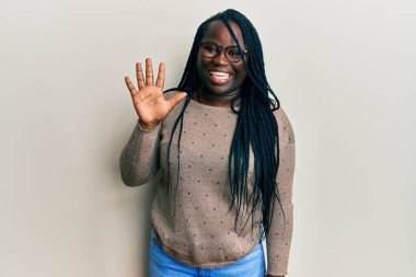 Saçları örgülü, günlük kıyafetler giyen, gözlüklü genç siyahi bir kadın beş numarayı gösterip işaret ederken kendinden emin ve mutlu bir şekilde gülümsüyor.. 