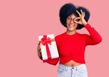 Genç Afrikalı Amerikalı kız elinde hediyeyle el hareketi yapıyor. Gülümsüyor, gözleriyle mutlu yüzle parmaklarının arasından bakıyor.. 