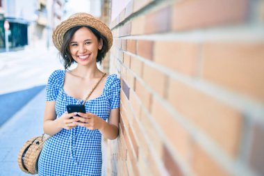 Genç ve güzel bir kız mutlu bir şekilde gülümsüyor. Akıllı telefon kullanıyor ve şehrin duvarına yaslanıyor.