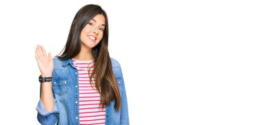 Genç esmer kadın gündelik kıyafetler giyip selam veriyor mutlu ve gülümsüyor, dostça karşılama hareketi yapıyor. 