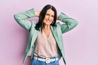 Orta yaşlı esmer kadın rahat kıyafetler giyiyor ve esniyor, kolları ve elleri başının arkasında mutlu bir şekilde gülümsüyor. 