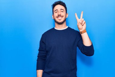 Gündelik kıyafetler giyen, gülümseyen ve kameraya göz kırpan genç İspanyol adam zafer işareti yapıyor. İki numara.. 