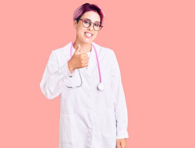 Pembe saçlı, doktor üniforması giyen genç ve güzel bir kadın elleriyle mutlu bir jest yapıyor. Kameraya bakıp başarıyı gösteren ifade. 