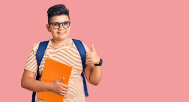 Sırt çantalı küçük bir çocuk elinde kitaplarla mutlu ve pozitif gülümsüyor, başparmağıyla mükemmel ve onay işareti yapıyor. 