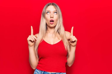 Gündelik kıyafetler giyen genç sarışın kadın şaşırmış ve şaşırmış bir şekilde yukarı bakıyor ve parmaklarıyla işaret ediyor ve kollarını kaldırıyor.. 