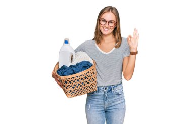 Çamaşır sepeti ve deterjan şişesi tutan güzel sarışın kadın parmaklarıyla imza atıyor, gülümseyerek mükemmel bir sembol oluşturuyor. 