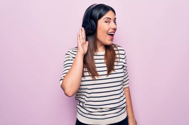Young beautiful brunette woman listening to music using headphones over pink background smiling with hand over ear listening and hearing to rumor or gossip. Deafness concept.
