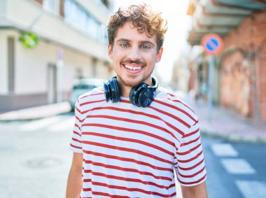 Genç beyaz adam mutlu bir şekilde gülümsüyor. Kulaklık takıyor ve şehirde yürüyor..