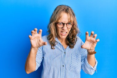 Orta yaşlı sarışın kadın günlük kıyafetler ve gözlük takıyor. Gülümsüyor. Kedi gibi pençe hareketi yapıyor, agresif ve seksi bir ifade. 