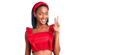 Genç Afro-Amerikan kadın günlük yaz kıyafetleri giyip kameraya gülümseyen ve zafer işareti yapan. İki numara.. 