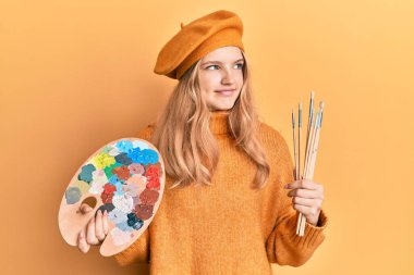 Güzel beyaz bir kız elinde boya fırçası ve ressam paleti ile yan tarafa bakıp düşünüyor.. 