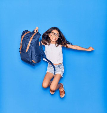 Sevimli İspanyol öğrenci kız gözlüklü mutlu gülümsüyor. Sırt çantasıyla sırtüstü sırtüstü uzanmış mavi arka plan