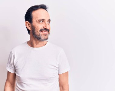 Orta yaşlı yakışıklı bir adam günlük tişört giyip izole edilmiş beyaz arka planda duruyor. Yan tarafa bakıyor, rahat bir görünüm takınıyor ve kendine güvenen bir gülümseme sergiliyor..