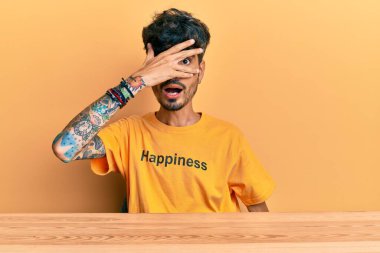 Üzerinde mutluluk mesajı olan İspanyol tişörtü giyen genç adam masaya oturmuş şok içinde yüzünü ve gözlerini kapayarak utanmış bir ifadeyle parmaklarının arasından bakıyor.. 