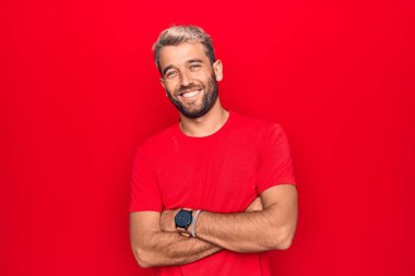 Genç yakışıklı, kırmızı tişört giyen, izole edilmiş kırmızı arka planda duran, kollarını kavuşturup kameraya bakan sarışın bir adam. Pozitif kişi.