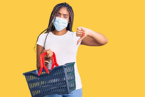 若いアフリカ系アメリカ人の女性は ショッピングバスケットと怒っている顔をした医療マスクを身に着けています 親指で嫌いを示す負の記号ダウン 拒絶概念 — ストック写真