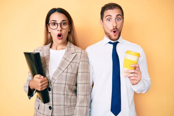 Beautiful couple wearing business clothes drinking take away coffee holding folder scared and amazed with open mouth for surprise, disbelief face