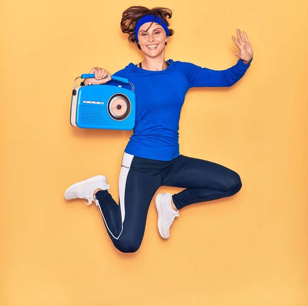 若い美しいスポーツ女性の音楽を聞いて幸せな笑顔 ジャンプとともに笑顔上の顔保持ヴィンテージラジオ上の隔離された黄色の背景 — ストック写真