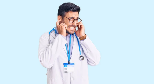 医者の制服を着た若いヒスパニック系の男と 大きな音楽のノイズのためにいらいらする表情で指で耳を覆う聴診器 聴覚障害の概念 — ストック写真