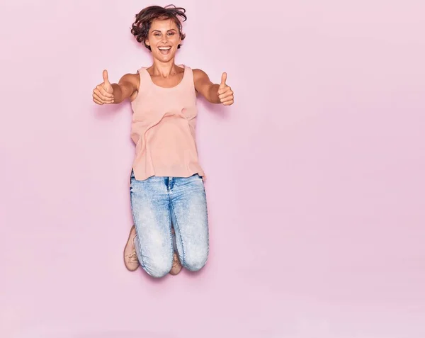 若い美しい女性が幸せな笑顔カジュアルな服を着て 顔に笑顔でジャンプします Signとともに親指アップ上の隔離されたピンクの背景 — ストック写真