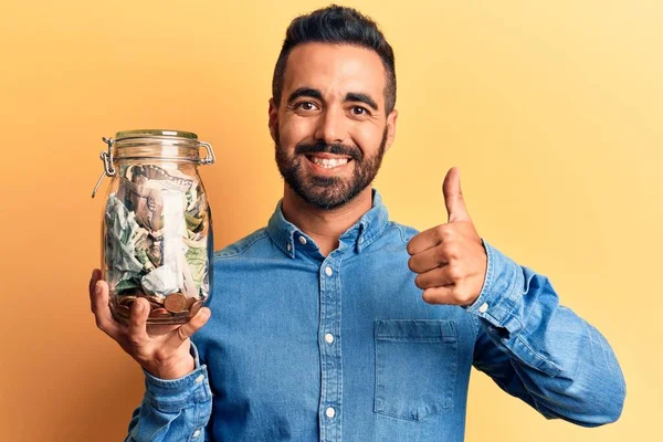 Молодой Латиноамериканец Держит Банку Сбережениями Улыбаясь Счастливым Позитивным Большой Палец — стоковое фото