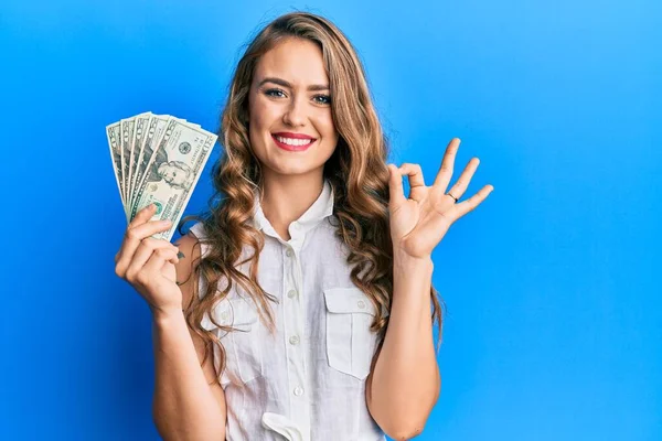 Young Blonde Girl Holding Dollars Doing Sign Fingers Smiling Friendly — Stock Photo, Image