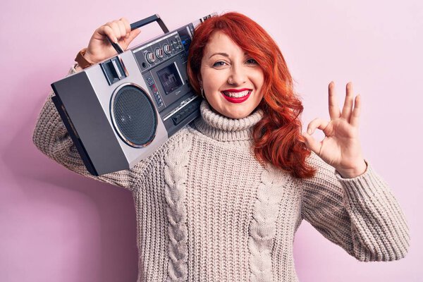 Beautiful redhead woman listening to music holding vintage boombox over pink background doing ok sign with fingers, smiling friendly gesturing excellent symbol