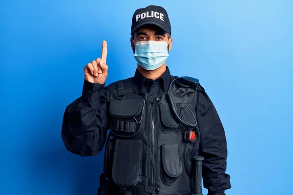 警察の制服を着た若いヒスパニック系の男と医療マスク笑顔でアイデアや質問を指差す指で幸せな顔 ナンバーワン — ストック写真