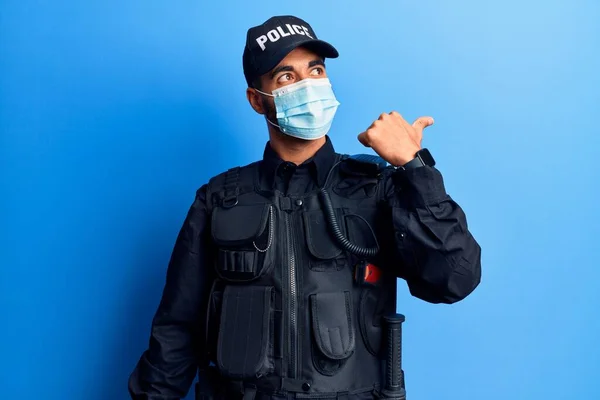 Young hispanic man wearing police uniform and medical mask pointing thumb up to the side smiling happy with open mouth