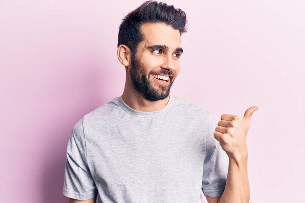 Young handsome man with beard wearing casual t-shirt pointing thumb up to the side smiling happy with open mouth 