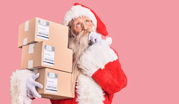 Old senior man with grey hair and long beard wearing santa claus costume holding boxes serious face thinking about question with hand on chin, thoughtful about confusing idea