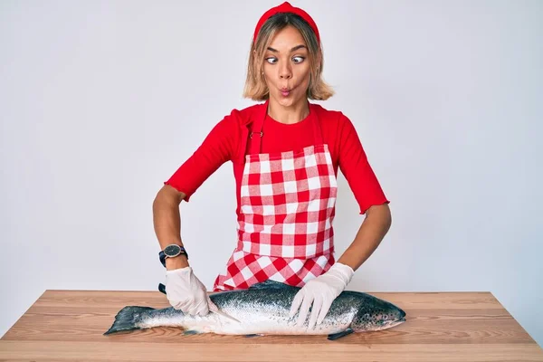 Beautiful caucasian woman fishmonger selling fresh raw salmon making fish face with mouth and squinting eyes, crazy and comical.