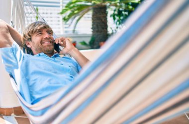 Orta yaşlı yakışıklı bir adam evinin terasında, bir hamakta uzanıp telefonda konuşuyor.