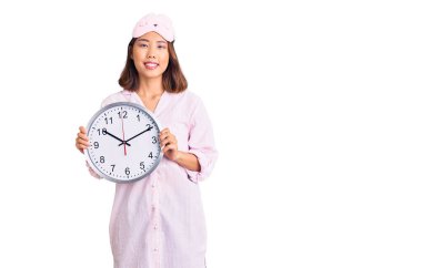 Uyku maskesi takan genç ve güzel bir Çinli kız ve elinde büyük bir saat tutan pijama. Pozitif ve mutlu görünüyor. Kendine güvenen bir gülümsemeyle dişlerini gösteriyor. 