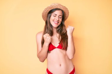 Bikini ve şapka giyen güzel beyaz kadın kolları havada ve gözleri kapalı bir şekilde başarıyı kutluyor ve şaşırıyor. 