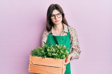 Genç, beyaz, bahçıvan önlüğü giyen genç bir kız elinde tahta bir saksı tutuyor şüpheci ve gergin, sorun yüzünden surat asıyor. negatif kişi. 