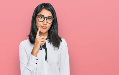 İş gömleği ve gözlük takan güzel Asyalı genç bir kadın ciddi bir yüz ifadesiyle eli çenesinde soru sorarken, kafa karıştırıcı bir fikir hakkında düşünceli. 