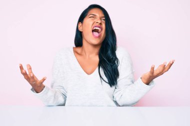 Uzun saçlı, gündelik giysiler giymiş güzel Latin bir kadın masaya oturmuş deli deli deli bağırıyor ve agresif bir ifadeyle bağırıyor ve kollarını kaldırıyor. hüsran kavramı. 