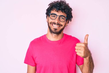 Kıvırcık saçlı, günlük kıyafetler giyen ve gözlüklü, mutlu ve pozitif gülümseyen, mükemmel ve onay işareti yapan yakışıklı bir genç. 