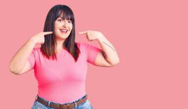 Genç ve cüsseli bir kadın günlük kıyafetler giyiyor neşeli bir şekilde gülümsüyor ve parmaklarıyla ve ağzıyla işaret ediyor. diş sağlığı kavramı. 