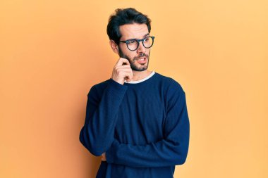 Gündelik kıyafetler ve gözlük takan genç İspanyol bir adam ciddi bir yüz ifadesi, eli çenesinde soru soran, kafa karıştırıcı bir fikir hakkında düşünceli. 