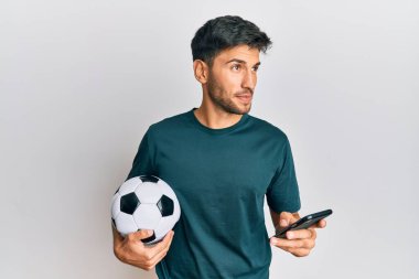 Genç yakışıklı adam futbol topu tutuyor. Akıllı telefon bahis uygulamasına bakıyor. Gülümsüyor ve başka tarafa bakıyor.. 