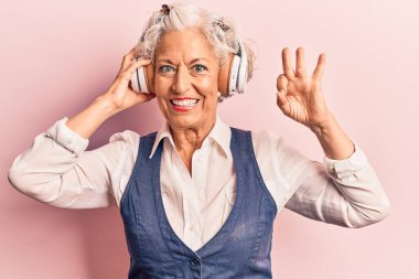 Yaşlı, kır saçlı, müzik dinleyen, kulaklık takan, parmaklarıyla imza atan, gülümseyen, dost canlısı, mükemmel bir sembol olan bir kadın. 