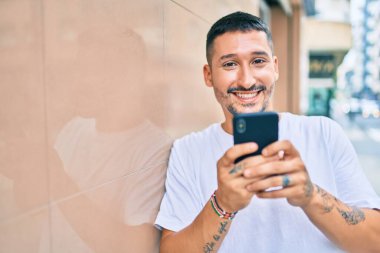 Duvara dayanmış akıllı telefon kullanan, mutlu gülümseyen genç İspanyol adam.