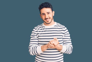 Latin asıllı genç adam ellerinin birleştiği günlük kıyafetler giyiyor ve gülümseyerek gevşemiş ve neşeli gülümsüyor. Başarı ve iyimserlik 