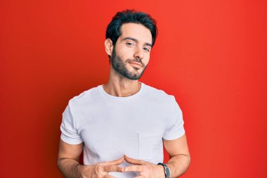 Latin kökenli genç adam, günlük beyaz tişörtler giyiyor ellerini birleştirip gülümseyerek gevşemiş ve neşeli olmasını diliyordu. Başarı ve iyimserlik 