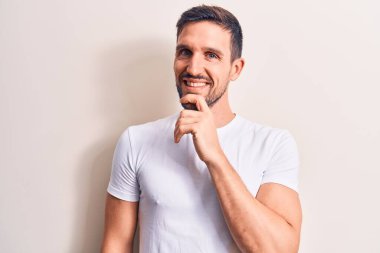 İzole edilmiş beyaz arka plan üzerinde duran rahat tişört giyen genç yakışıklı adam kameraya çapraz kollar ve çenesi üzerinde kendinden emin bir şekilde bakıyor. Olumlu düşünüyorum..